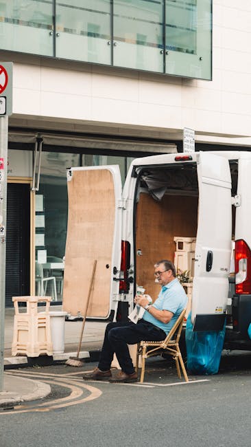 A man wearing glasses and a light blue shirt is seated on a wooden chair outside a white delivery van parked on the street in an urban area. The van's rear doors are open, revealing the interior filled with flat-packed furniture, cardboard boxes, and wrapped items prepared for home relocation. The man appears to be reading or checking a document while resting, with packing materials like foam wrap and plastic covering visible among the objects inside the van. Nearby, a broom is propped against a wooden panel, and a large wooden board leans against the open van door, indicating a loading or unloading process. The setting includes a modern building with glass windows and a no-parking sign on the street, with the scene captured in daylight under natural lighting, reflecting the logistical aspects of furniture transport and packing for removals services as offered by Man with Van Kensington.