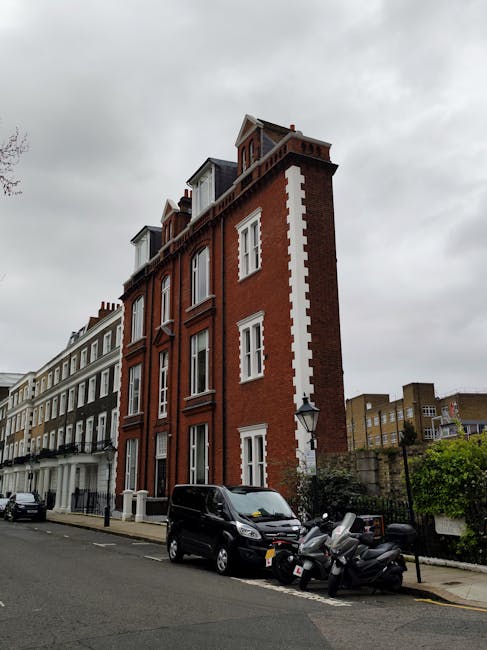 A multi-storey brick building with a red façade and white decorative details on the corners located on a residential street under a cloudy sky. The building features several large sash windows, some with white frames, and has multiple small dormer windows on the top floor. In the foreground, a black van is parked on the street alongside three motorcycles. The street is lined with other residential properties, and a lamppost is positioned near the foreground. This image illustrates an urban setting suitable for house removals and home relocation services, with ample space for loading and unloading goods, consistent with the operations of Man with Van Kensington involved in furniture transport and packing and moving activities.