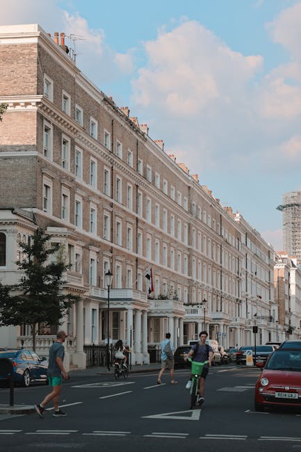 A multi-storey brick building with a red façade and white decorative details on the corners located on a residential street under a cloudy sky. The building features several large sash windows, some with white frames, and has multiple small dormer windows on the top floor. In the foreground, a black van is parked on the street alongside three motorcycles. The street is lined with other residential properties, and a lamppost is positioned near the foreground. This image illustrates an urban setting suitable for house removals and home relocation services, with ample space for loading and unloading goods, consistent with the operations of Man with Van Kensington involved in furniture transport and packing and moving activities.