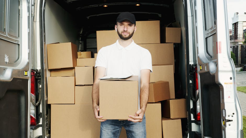 A young man wearing a white shirt, dark cap, and jeans stands inside the open rear doors of a white moving van, holding a cardboard box in his hands. The back of the van is filled with numerous stacked cardboard boxes of various sizes, some wrapped in plastic or packing paper, indicating a home relocation process. The boxes are arranged to maximize space within the vehicle, with some placed on their sides for stability. The van is parked on a residential street with houses visible in the background, and bright natural daylight illuminates the scene. The man appears to be involved in furniture transport or packing and loading tasks, aligning with house removals services provided by Man with Van Kensington. Equipment such as trolleys, straps, or blankets are not visible, but the setting emphasizes the logistics of moving items securely and efficiently during a furniture transport or packing and moving operation.