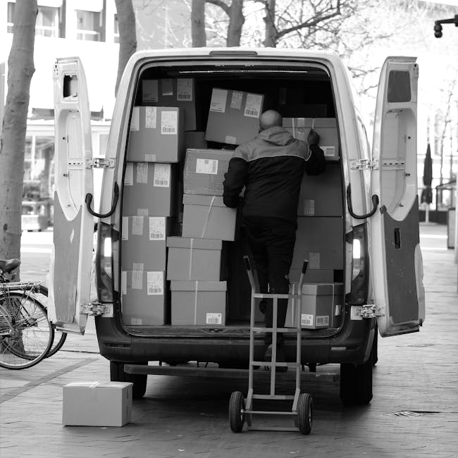 A man wearing a jacket is standing inside the open back of a white delivery van, surrounded by several stacked cardboard boxes of varying sizes, secured with labels for identification. He is handling one of the boxes as part of a packing and loading process for a house removal. The van, parked on a paved street outdoors, has its rear doors swung open, revealing the interior filled with more boxes arranged to maximize space. A small hand trolley is positioned nearby, ready to assist with transporting the packed items. In the background, there are bicycles parked along the sidewalk and buildings with large windows, indicating an urban environment. Natural daylight illuminates the scene, capturing the logistics involved in furniture transport and home relocation services. This image exemplifies professional moving operations by Man with Van Kensington, emphasizing careful packing, efficient loading, and the preparation required for home removals within a city setting.