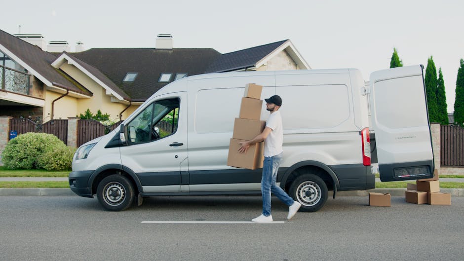 A young man wearing a white shirt, dark cap, and jeans stands inside the open rear doors of a white moving van, holding a cardboard box in his hands. The back of the van is filled with numerous stacked cardboard boxes of various sizes, some wrapped in plastic or packing paper, indicating a home relocation process. The boxes are arranged to maximize space within the vehicle, with some placed on their sides for stability. The van is parked on a residential street with houses visible in the background, and bright natural daylight illuminates the scene. The man appears to be involved in furniture transport or packing and loading tasks, aligning with house removals services provided by Man with Van Kensington. Equipment such as trolleys, straps, or blankets are not visible, but the setting emphasizes the logistics of moving items securely and efficiently during a furniture transport or packing and moving operation.