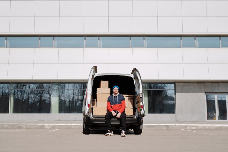 A man wearing a red and blue jacket and a blue beanie hat stands outdoors next to a silver delivery van with its rear doors open. Inside the van, stacked cardboard boxes and packing materials are visi