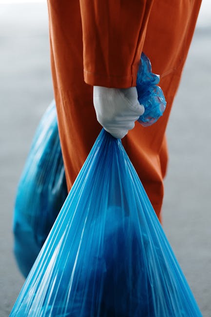 A person dressed in an orange high-visibility work suit, wearing a white glove, is holding a large blue plastic disposal bag filled with waste. The individual is standing outdoors on a paved surface, 
