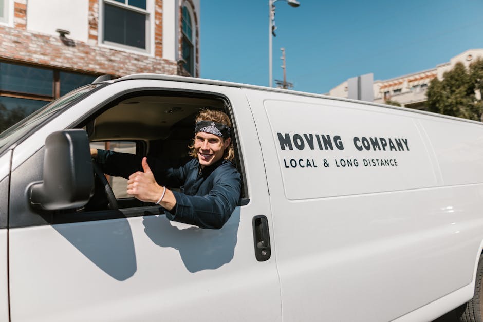 A young male mover with light brown, wavy hair wearing a black bandana and navy blue work shirt sits in the driver's seat of a white van, giving a thumbs-up and smiling. The van has the text 'MOVING C