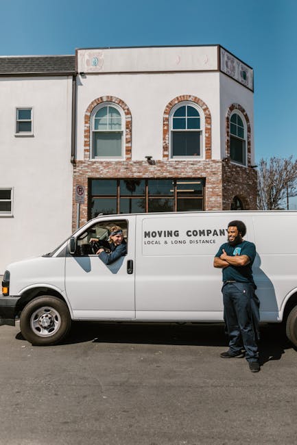 A young male mover with light brown, wavy hair wearing a black bandana and navy blue work shirt sits in the driver's seat of a white van, giving a thumbs-up and smiling. The van has the text 'MOVING C