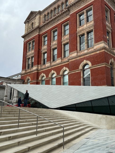 The image shows the exterior of a historic red brick building with arched windows and detailed stonework, situated beneath a cloudy sky. A modern, angular glass structure extends from the building, with steps leading up to it. On the steps, two individuals are visible, one standing and the other walking, both wearing dark clothing. The scene is part of a building entrance or outdoor area connected to the London museum, likely involved in house removal or moving activities with Man with Van Kensington. The setting indicates a professional relocation process, with the individuals possibly preparing to load or unload items, in an environment designed for efficient furniture transport or packing during a home relocation.