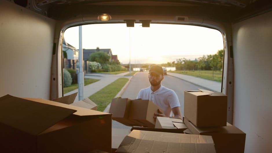 A man with a beard, wearing a white polo shirt and sunglasses, is seen loading cardboard boxes into the back of a moving van during a home relocation. The van's interior is visible, with several seale