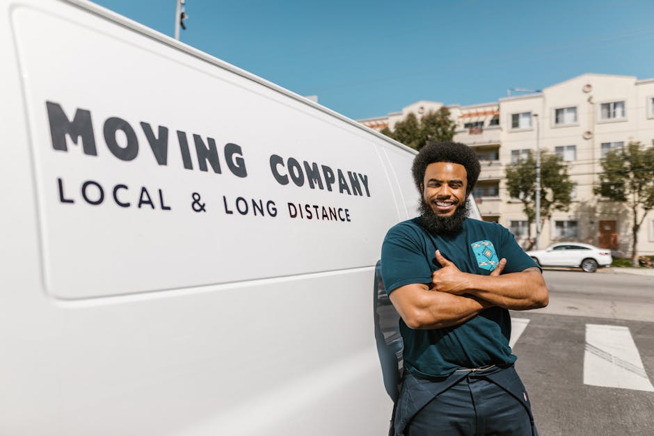 A man with a beard, wearing a white polo shirt and sunglasses, is seen loading cardboard boxes into the back of a moving van during a home relocation. The van's interior is visible, with several seale