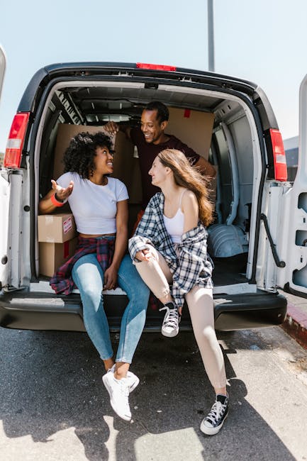 Two young women sit inside the open rear of a moving van parked on a paved driveway, engaged in conversation and smiling at each other. One woman has curly black hair, wearing a light blue T-shirt, and is gesturing with her hand. The other has long, wavy brown hair, wearing a white top and a plaid shirt tied around her waist. Behind them, a man is leaning inside the van, smiling and looking at the women. The interior of the van contains cardboard boxes, some wrapped in plastic or packing paper, and plastic storage bins. The van's service area shows a spacious cargo space with visible ceiling railings and tie-down points, suitable for home relocation and furniture transport. The scene suggests careful packing and loading preparations for a house move, with the vehicle designed for transporting household items during a furniture moving process, illustrating the logistics of packing and loading during a house removal.