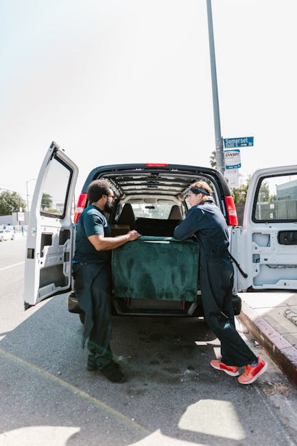 Two professional movers from Man with Van Kensington are engaged in a home relocation process, loading a large, green upholstered piece of furniture into the open cargo area of a black van parked on a street. One mover is using lifting straps to support the furniture, while the other holds the piece steady and guides it into the vehicle. The interior of the van is visible, revealing a spacious area suited for transporting household items. The scene is set outdoors on a sunny day, with a clear sky and a street sign visible in the background. The surrounding environment includes pavement, a sidewalk with a pink curb, and some distant commercial buildings, emphasizing the logistics involved in furniture transport and packing during a house move. The professional team, part of Man with Van Kensington, is demonstrating careful handling and efficient loading as part of their removals service.