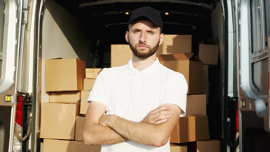 A young man with a beard and short dark hair, dressed in a white polo shirt and a black cap, stands with arms crossed inside the open rear of a moving van filled with neatly stacked cardboard moving boxes of various sizes. The van's interior is well-lit and visible behind him, showing an organized loading process with some furniture covered in protective fabric or plastic wrap. Outside the van, the surrounding environment appears to be a residential area, with pavement visible at the bottom of the image. The scene depicts a professional home relocation or furniture transport setting, relevant to house removals and packing services offered by Man with Van Kensington, as seen on the webpage titled 'W8 to SW7 move price comparison man with van rates'.