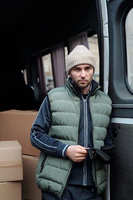 A man wearing a white polo shirt, black cap, and white earbuds is inside the open back of a large van, which is packed with numerous cardboard boxes of varying sizes. He is taking notes on a clipboard while standing beside a woman with long red hair, dressed in a black sleeveless top, light blue jeans, and a white cap, who is holding a cardboard box. The van is situated on a paved area outside a building, with sunlight illuminating the scene, as the moving process involves loading or unloading boxes during a home relocation. The interior of the van shows a neatly stacked arrangement of boxes, some secured with plastic wrap, and a top-down view captures the space used for furniture transport and packing during a professional removal service. Man with Van Kensington's crew appears to be systematically organizing items for efficient moving and logistics, reflecting typical packing and loading activities involved in house removals.
