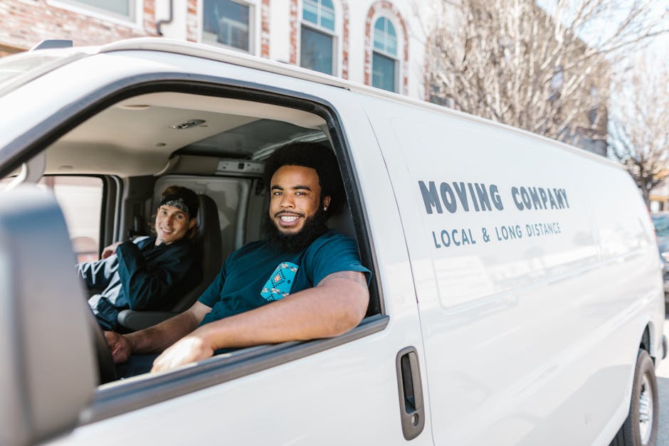 A white moving van with the words 'MOVING COMPANY, LOCAL & LONG DISTANCE' printed on its side is parked on a street in Kensington. Inside the driver's cabin, a smiling man with dark curly hair and a beard is seated in the front, wearing a dark T-shirt with a partially visible logo, and appears to be engaging with the camera. Next to him, a woman with dark hair tied back, wearing a black jacket and a headband, is seated in the passenger seat, also smiling. The background shows residential buildings with large windows, trees, and a clear sky, indicating a daytime home relocation or furniture transport process. The van is positioned next to a pavement, suggesting it is in the process of loading or unloading household items, with packing materials or furniture potentially inside. This image reflects a professional moving service, such as that offered by Man with Van Kensington, involved in the logistics of house removals and packing during a home move.