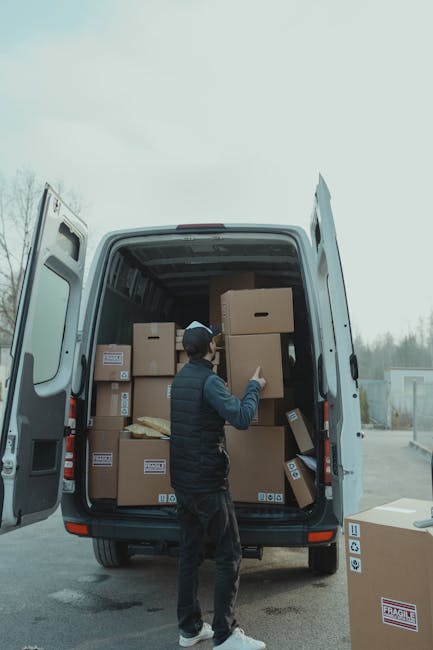 A man dressed in dark clothing, including a cap, is standing outside on a paved area in front of a large white delivery van with its rear doors wide open. Inside the van, numerous cardboard boxes of various sizes are stacked, some with visible labels, with a few boxes placed on top of others, indicating an active loading process typical of house removals or home relocation tasks. The man is seen lifting or moving one of the medium-sized boxes towards the interior of the van. Additional boxes are positioned on the pavement nearby, ready to be transported, with some partially visible in the foreground. The environment appears to be an outdoor loading area, possibly in a residential or commercial setting, with overcast weather and a slightly blurred background of trees and industrial structures. This image features elements such as packing materials, cardboard boxes, a moving van, and loading equipment, accurately depicting aspects of furniture transport, packing, and moving logistics carried out by professional removal companies like Man with Van Kensington.